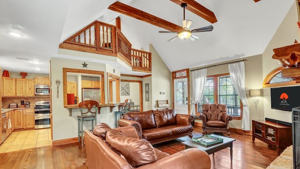 A spacious living room with leather sofas, a wooden staircase, a kitchen with wooden cabinets, and a ceiling fan. - Secluded Cabin for 10 guests with Pool, Hot Tub, and Fire Pit (Wimberley)