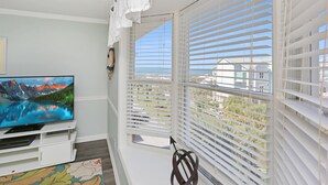 A balcony with white blinds, a mounted TV, and a view of a mountain lake. - Gated Community, Pool, Green Space, Great Beach Access - SeaCliff (Cape San Blas)