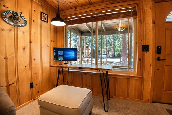 Un intérieur en bois avec un bureau d’ordinateur, une fenêtre donnant sur une cabane et un panneau fixé au mur.