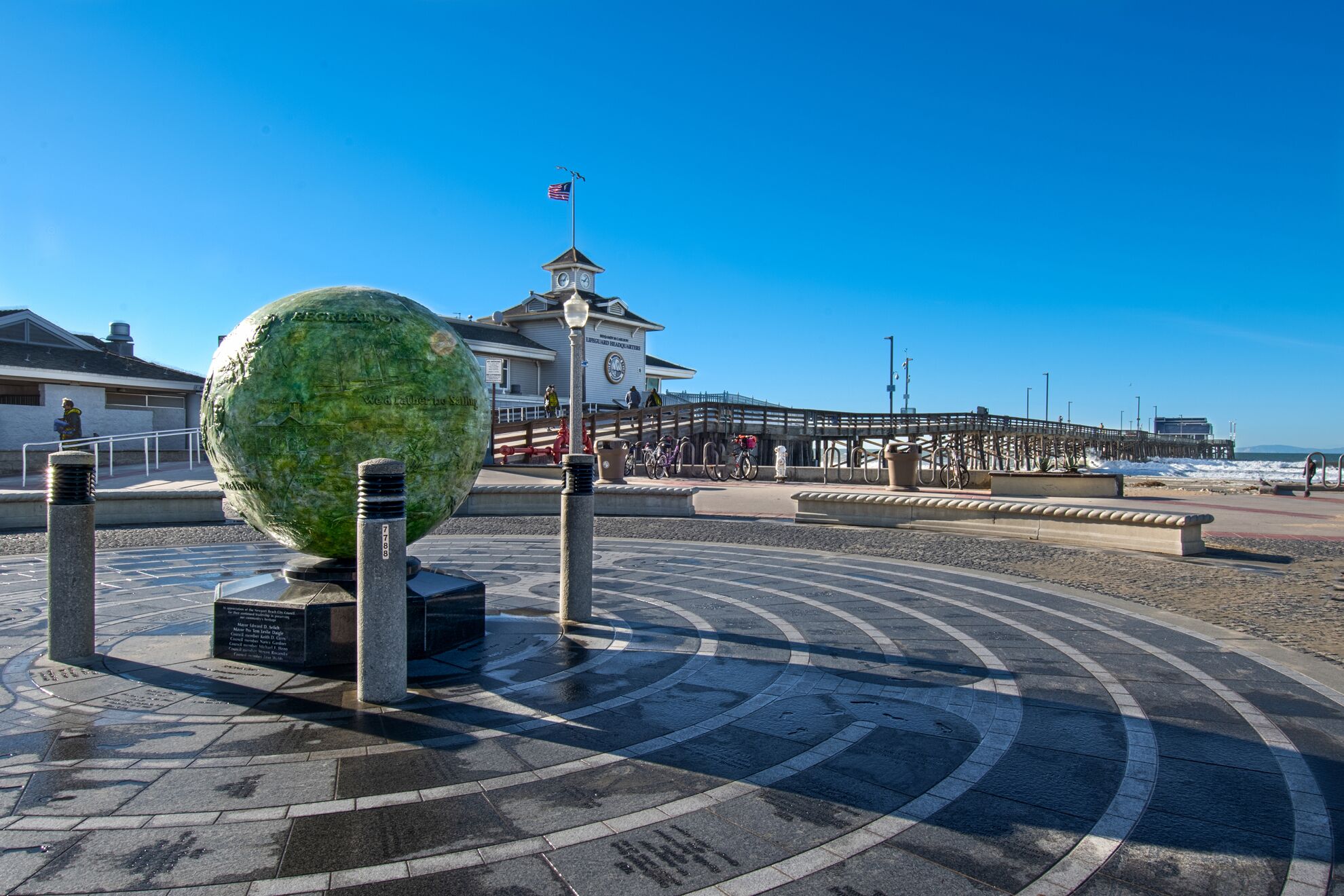 A waterfront promenade with a large green sculpture, a clock tower, and a pier with bicycles.