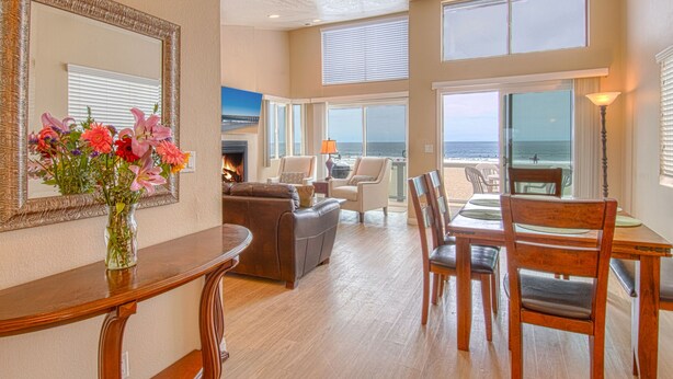 A spacious living room with a fireplace, a dining area with wooden furniture, and large windows offering ocean views. - Cheery and bright upstairs unit is a perfect beach getaway! (Newport Beach)