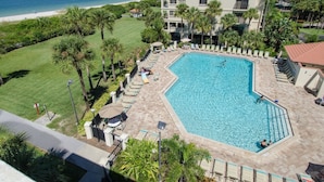 A swimming pool with lounge chairs and umbrellas, surrounded by palm trees and a view of the beach and ocean.