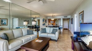 A spacious living room with a large sofa, a coffee table, and a TV. There is a kitchen in the background with a dining area.