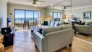 A spacious living room with a large sofa, a coffee table, and a TV. There is a balcony with a view of the ocean.