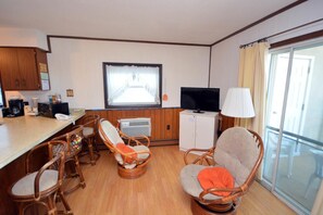 Habitación de hotel compacta con kitchenette, televisión, sillas de mimbre y un ventanal grande.
