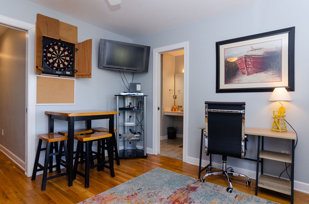 A room with a dartboard, TV, framed artwork, and a desk with a chair.