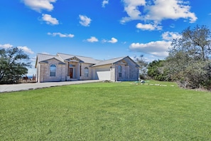 A two-story stone house with a front garage, a well-maintained lawn, and a clear blue sky with clouds. - Soak in only the BEST Hill Country views form your own private pool! (Wimberley)