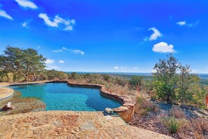Une piscine d’un bleu éclatant, bordée d’une terrasse en pierre, entourée d’arbres et offrant une vue sur le paysage.