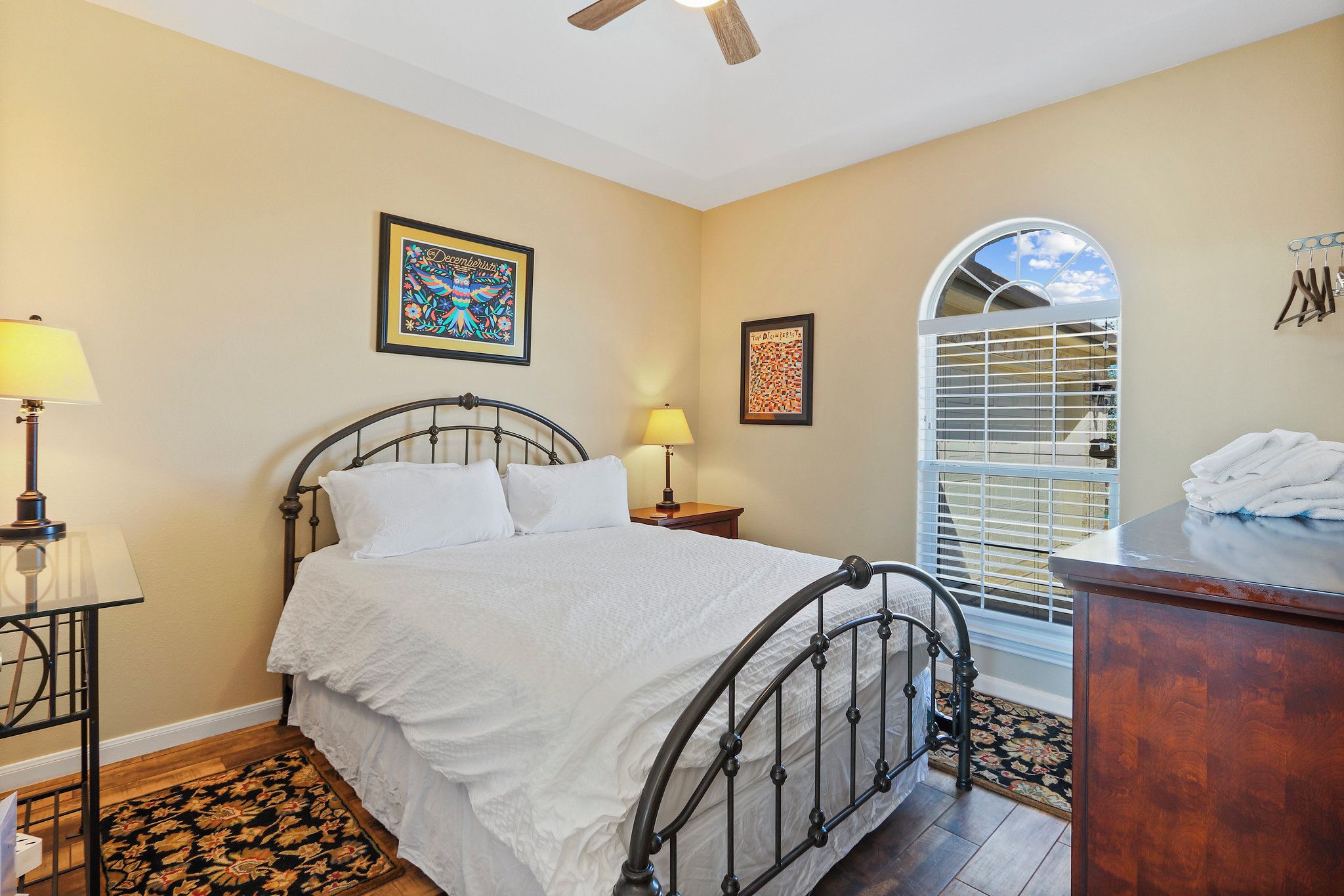 A bedroom with a bed, two bedside lamps, a wooden nightstand, a dresser, and a window with blinds.