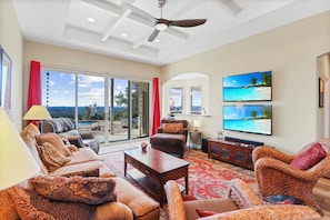 A spacious living room with a large window, a ceiling fan, and a flat-screen TV mounted on the wall. - Soak in only the BEST Hill Country views form your own private pool! (Wimberley)