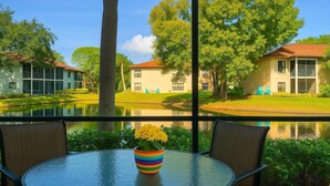 A view from a room with a large window overlooking a courtyard with a pond, trees, and buildings. There is a table with a colorful vase in the foreground. - Beautiful 2/2 ground floor condo ⚓ resort style community (Bradenton)