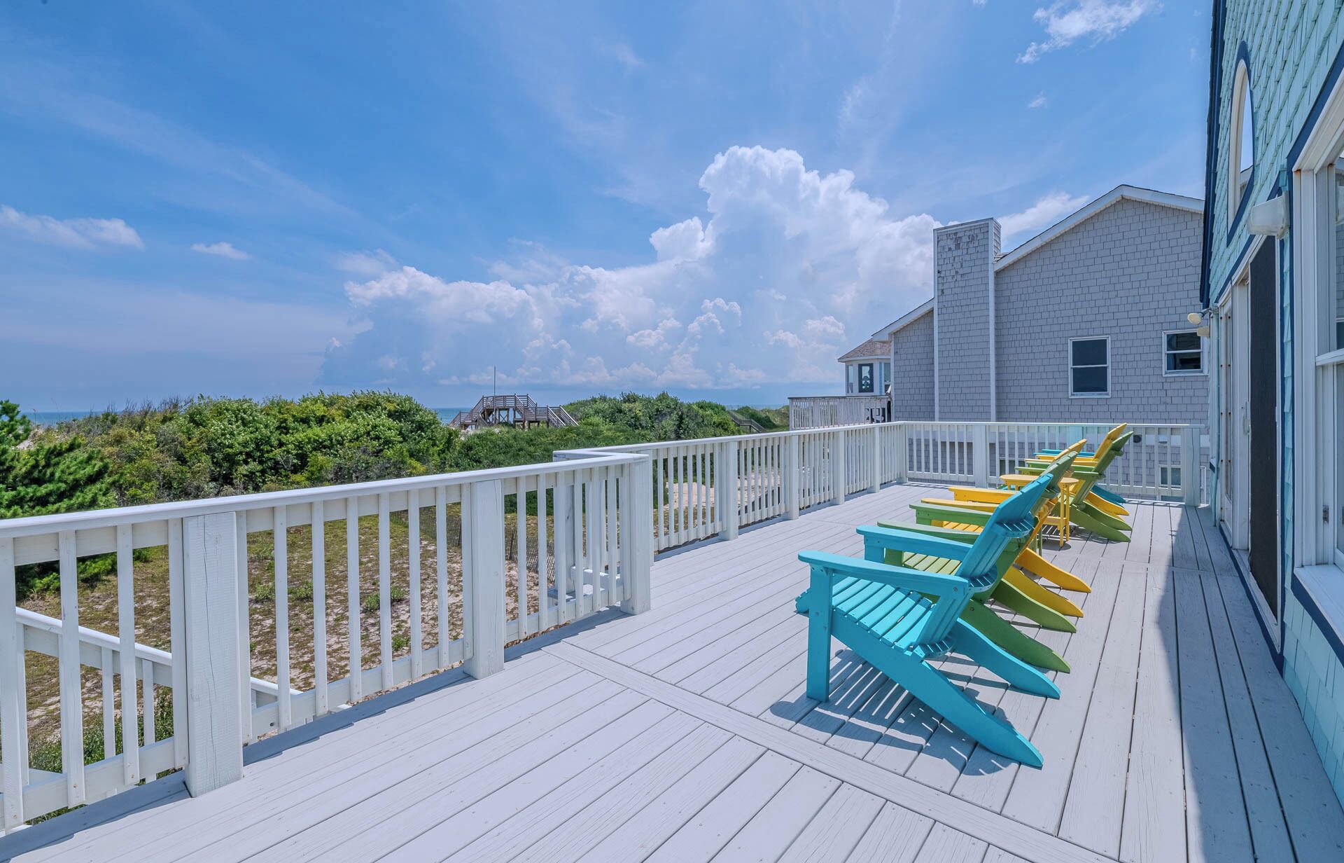 A wooden deck with a white railing, a row of colorful Adirondack chairs, and a building with a gray facade in the background.