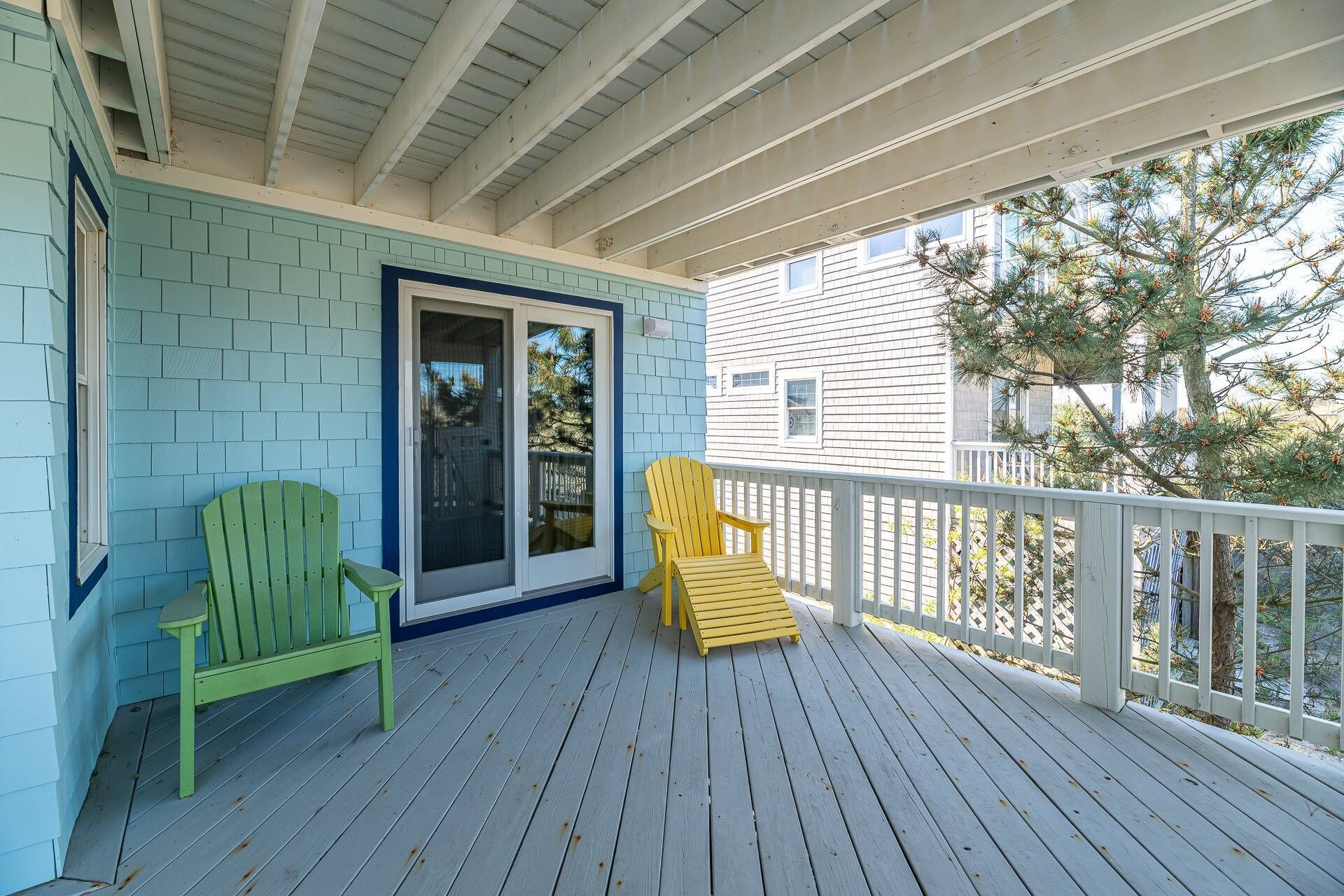 A covered porch with two Adirondack chairs, a sliding glass door, and a view of a neighboring house.