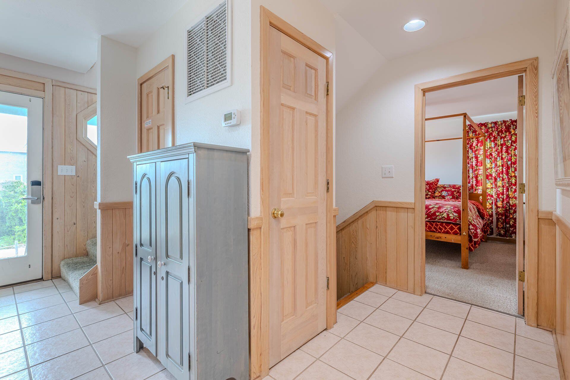 A hallway with a wooden wardrobe, a door leading to a bedroom with a bed and floral curtains, and a vent on the wall.