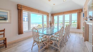 A spacious room with a glass-top dining table surrounded by wicker chairs, large windows with ocean views, and a ceiling with recessed lighting. - Terning Point - Corolla, Crown Point, Oceanfront, 20 Yds to Beach, Pool, Hot Tub, Pets OK (Corolla)