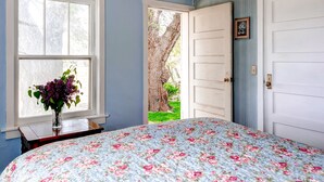 Ein Schlafzimmer mit floralem Bettbezug, einem Fenster mit Blumenvase und einer Tür, die in einen Garten führt.