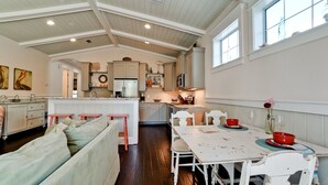 Una cocina amplia con techos altos y abundante luz natural, que cuenta con una isla central, electrodomésticos modernos y un rincón comedor acogedor con una mesa blanca y detalles en rojo.
