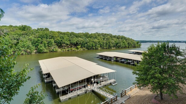 A lakeside property with multiple docked boats, a covered deck area, and a forested shoreline. - Tranquil Osage Beach Condo w/ Waterfront Decks! (Osage Beach)