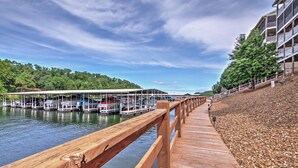 A wooden dock leading to a marina with boats docked, a covered boat launch, and a building with balconies on a hillside. - Tranquil Osage Beach Condo w/ Waterfront Decks! (Osage Beach)