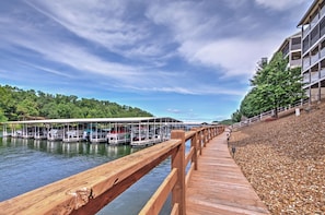A wooden dock leading to a marina with boats docked, a covered boat launch, and a building with balconies on a hillside. - Tranquil Osage Beach Condo w/ Waterfront Decks! (Osage Beach)
