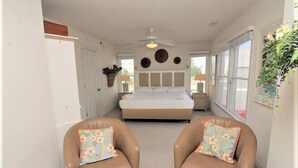 Un dormitorio con cama, dos sillas, ventilador de techo y una ventana con cortinas.