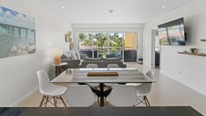 Een moderne woonkamer met een glazen eettafel, witte stoelen en een bank met kussens. Er is een tv aan de muur en een schilderij.