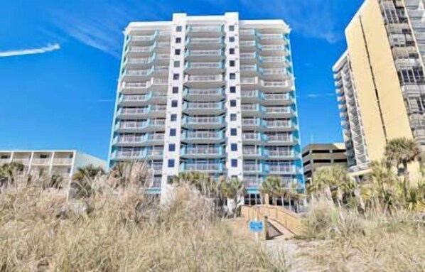 Un hotel de gran altura con múltiples balcones, situado cerca de una playa con vegetación escasa.