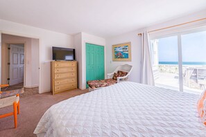 A bedroom with a large bed, a television on a dresser, a chair, and a view of the ocean through a large window. - Summer Camp - Corolla, Crown Point, Oceanfront, 70 Ft to Beach, Community Pool, Hot Tub, Pets OK (Corolla)