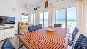 A room with a wooden table, a TV, and a sofa with cushions. There are windows with curtains and a view of the beach. - Summer Camp - Corolla, Crown Point, Oceanfront, 70 Ft to Beach, Community Pool, Hot Tub, Pets OK (Corolla)