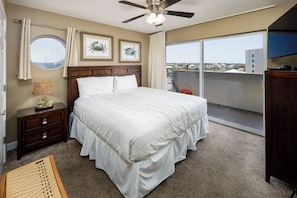 A bedroom with a large bed, a ceiling fan, a nightstand, a lamp, and a view of the city through a balcony.