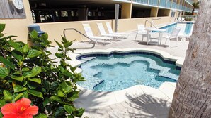 Un espace piscine doté de chaises longues et d’un jacuzzi, entouré de verdure et bordé d’un bâtiment avec des balcons.