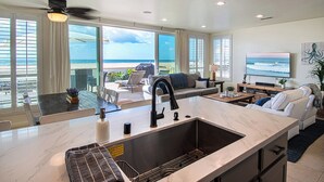 A modern kitchen with a large island, a dining area with a table and chairs, a living room with a sofa and TV, and large windows with a view of the beach. - Lovely lower unit with spectacular oversized patio right on the sand! (Newport Beach)