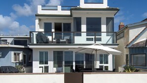 A two-story beachfront house with a balcony, large windows, and a covered outdoor seating area. - Lovely lower unit with spectacular oversized patio right on the sand! (Newport Beach)