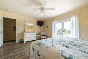 Un dormitorio con cama, ventilador de techo, cómoda, silla, televisor y balcón con cortinas.