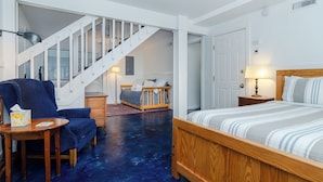 Quarto com cama de madeira, poltrona azul, mesa pequena, mesa de cabeceira e escada que leva a outro espaço.