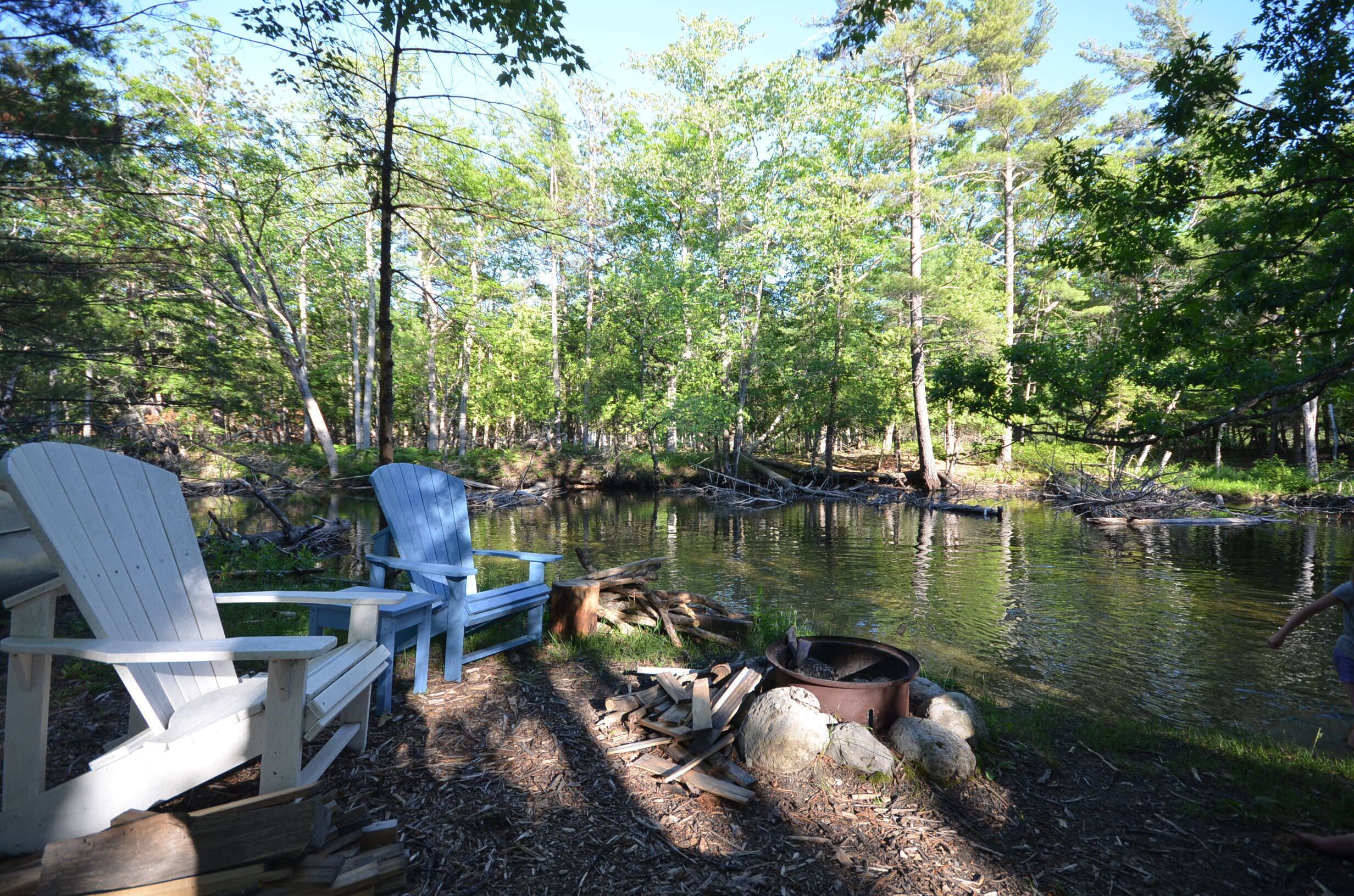 Ein Campingplatz am Seeufer mit Adirondack-Stühlen, einer Feuerstelle und einer rustikalen Umgebung, umgeben von Bäumen.