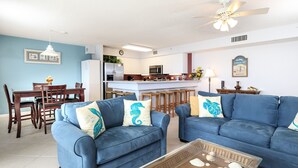A living room with two blue sofas, a coffee table with seashells, a dining area with wooden chairs, and a kitchen in the background.