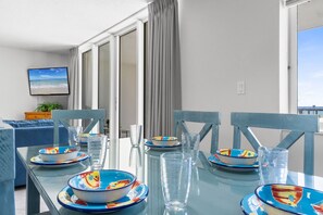 Un comedor moderno con una mesa de vidrio dispuesta para seis personas, sillas azules y vistas al mar a través de amplios ventanales.