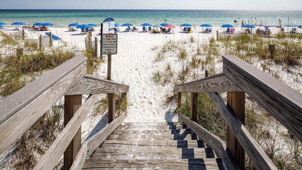 A wooden staircase leading to a sandy beach with blue umbrellas and beach chairs.