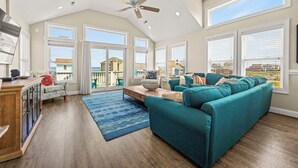 Amplio salón con un sofá grande color turquesa, una mesa de centro de madera y un tapete azul estampado. Hay un ventilador de techo, ventanas grandes con persianas y vistas a una playa y casas.