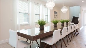 A modern dining area with a glass table, white chairs, potted plants, and a staircase in the background. - Stylish Sleek Home with Full Kitchen at Encore (Kissimmee)
