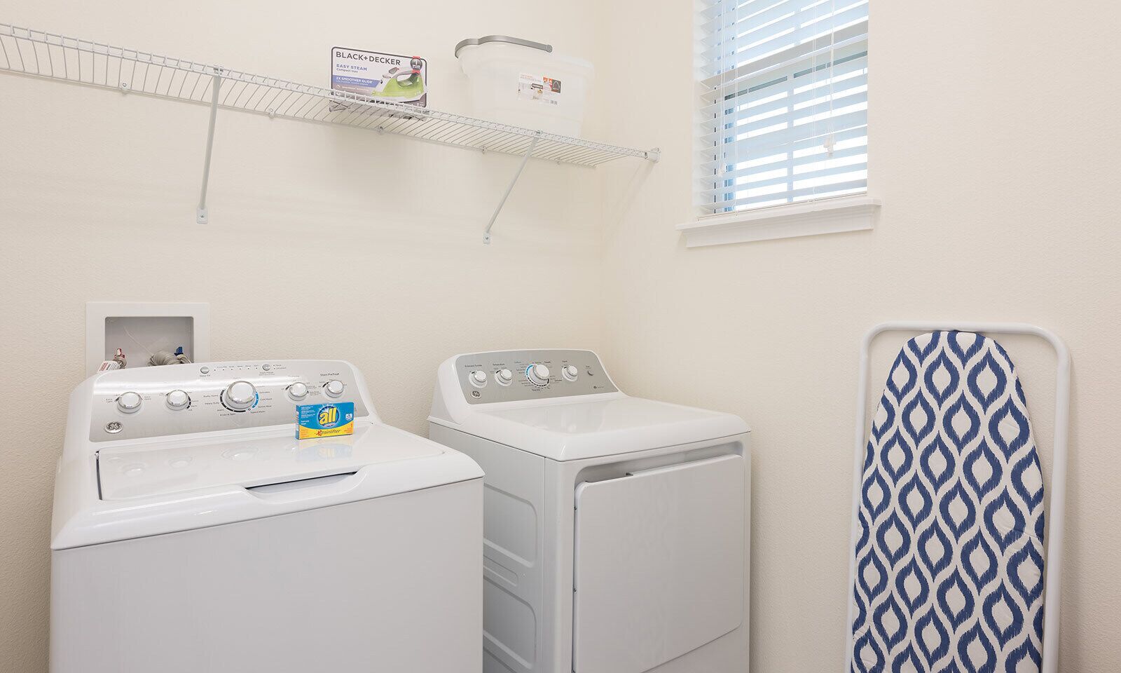 A laundry room with two white washing machines, a drying rack, and a shelf with a detergent box.