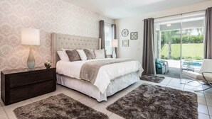 A bedroom with a large bed, a nightstand, a lamp, and a view of the outdoors through a sliding glass door. - Stylish Sleek Home with Full Kitchen at Encore (Kissimmee)