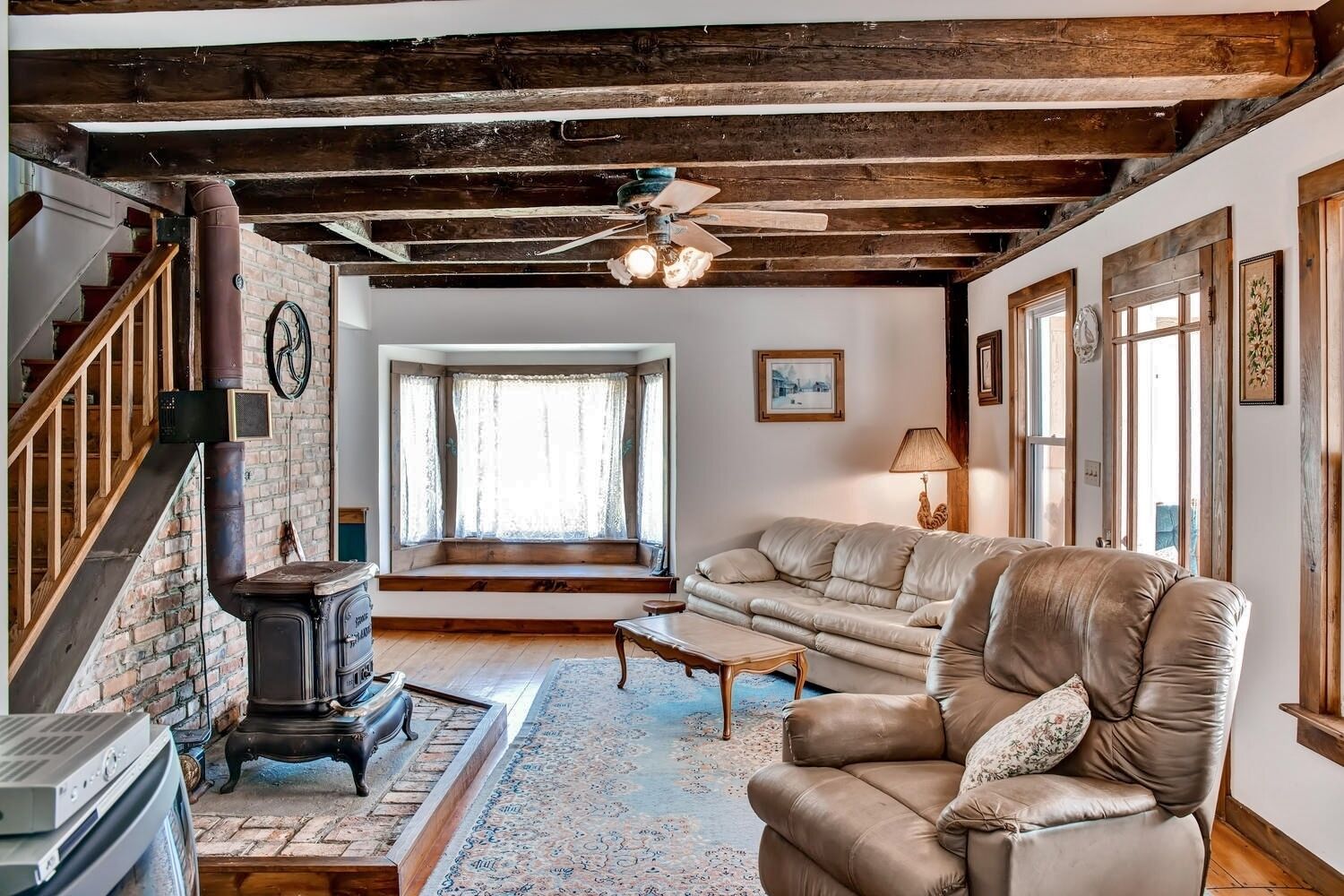 A cozy living room with exposed wooden beams, a leather sofa, a fireplace, and a staircase.