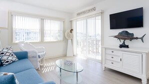 Un salon lumineux avec un canapé bleu, une table basse en verre, un meuble blanc et un téléviseur fixé au mur.