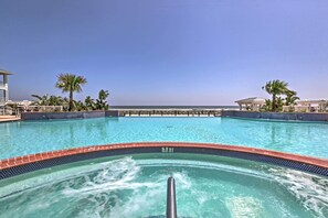 A clear blue pool with a 3 FT depth marker, surrounded by palm trees and lounge chairs, with a view of the ocean and a building in the background. - Steps to Ocean! Breezy Galveston House w/ 2 Decks (Galveston)