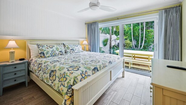 Une chambre à coucher comprenant un lit, des tables de chevet, un ventilateur de plafond, une fenêtre avec des rideaux et une vue sur la verdure à l’extérieur.