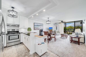 A modern kitchen with white cabinetry, a built-in oven, and a dining area with a table and chairs. - Epic West Maui View! Laundry, WiFi, TVs, Kitchen, AC, Ceiling Fans–Kamaole Sands 10202 (KIHEI)