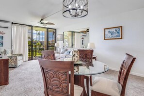 A spacious living room with a glass dining table, wooden chairs, a chandelier, and a view of the outdoors. - Epic West Maui View! Laundry, WiFi, TVs, Kitchen, AC, Ceiling Fans–Kamaole Sands 10202 (KIHEI)
