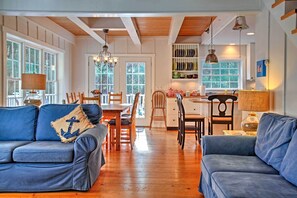 A spacious living area with a wooden floor, a blue sofa with an anchor pillow, a dining table with chairs, and a kitchen area with shelves and a hanging lamp.